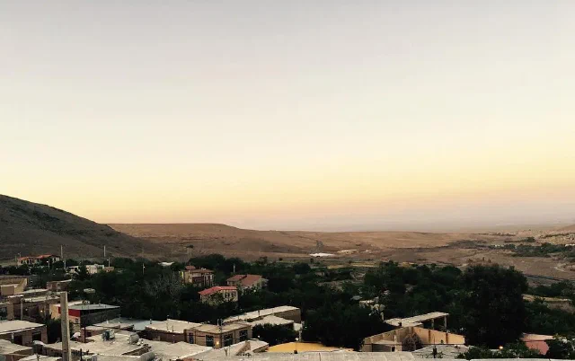 بام‌ها و درختان روستا زیر آسمان زرد روشن با تپه‌ها در دوردست