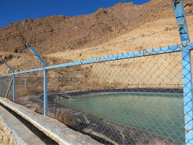 استخر ذخیره آب پشت حصار توری آبی با دامنه‌ای سنگی در پس‌زمینه